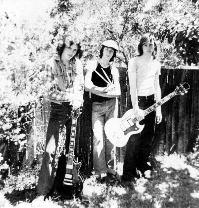 Three teenage musicians standing in a suburban backyard: one holding an electric guitar, one holding drumsticks, one holding a bass guitar.