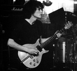 Black and white photo of a guitarist playing in front of a Marshall amplifier.
